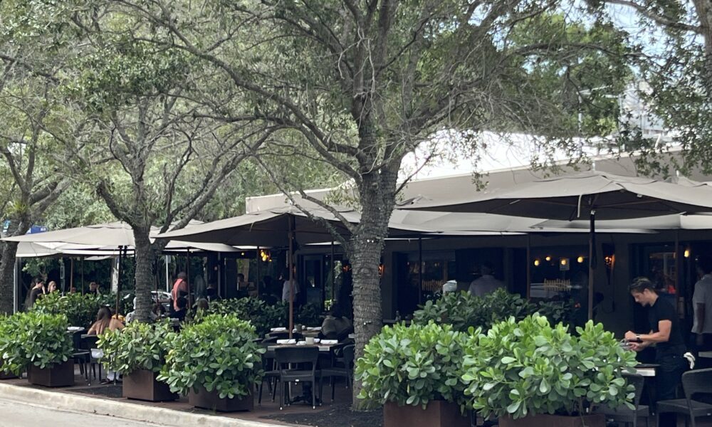 Commodore Plaza sidewalk cafe tables under tree canopy in Coconut Grove